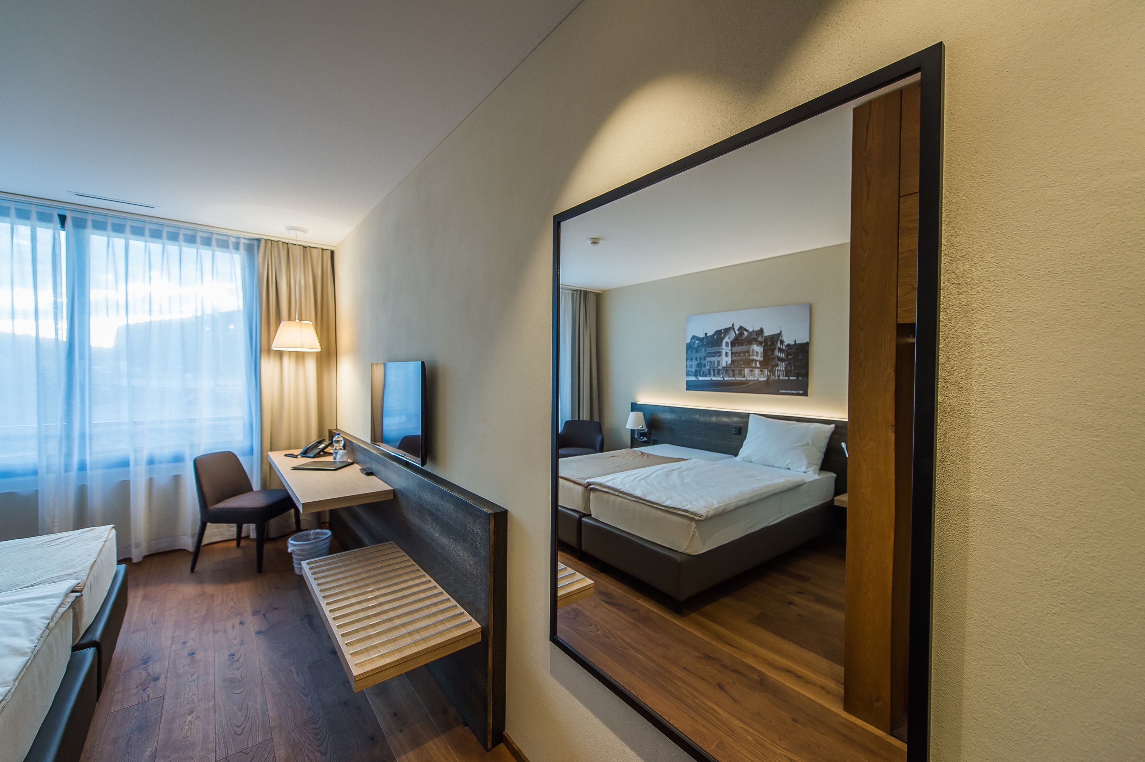 Une chambre d’hôtel comprenant un lit, un bureau, une chaise, une petite table, une lampe et un grand miroir.