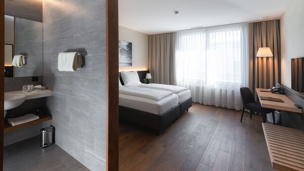 A modern hotel room with a large bed, a desk, a chair, a bathroom with a sink and mirror, and a large window with curtains.