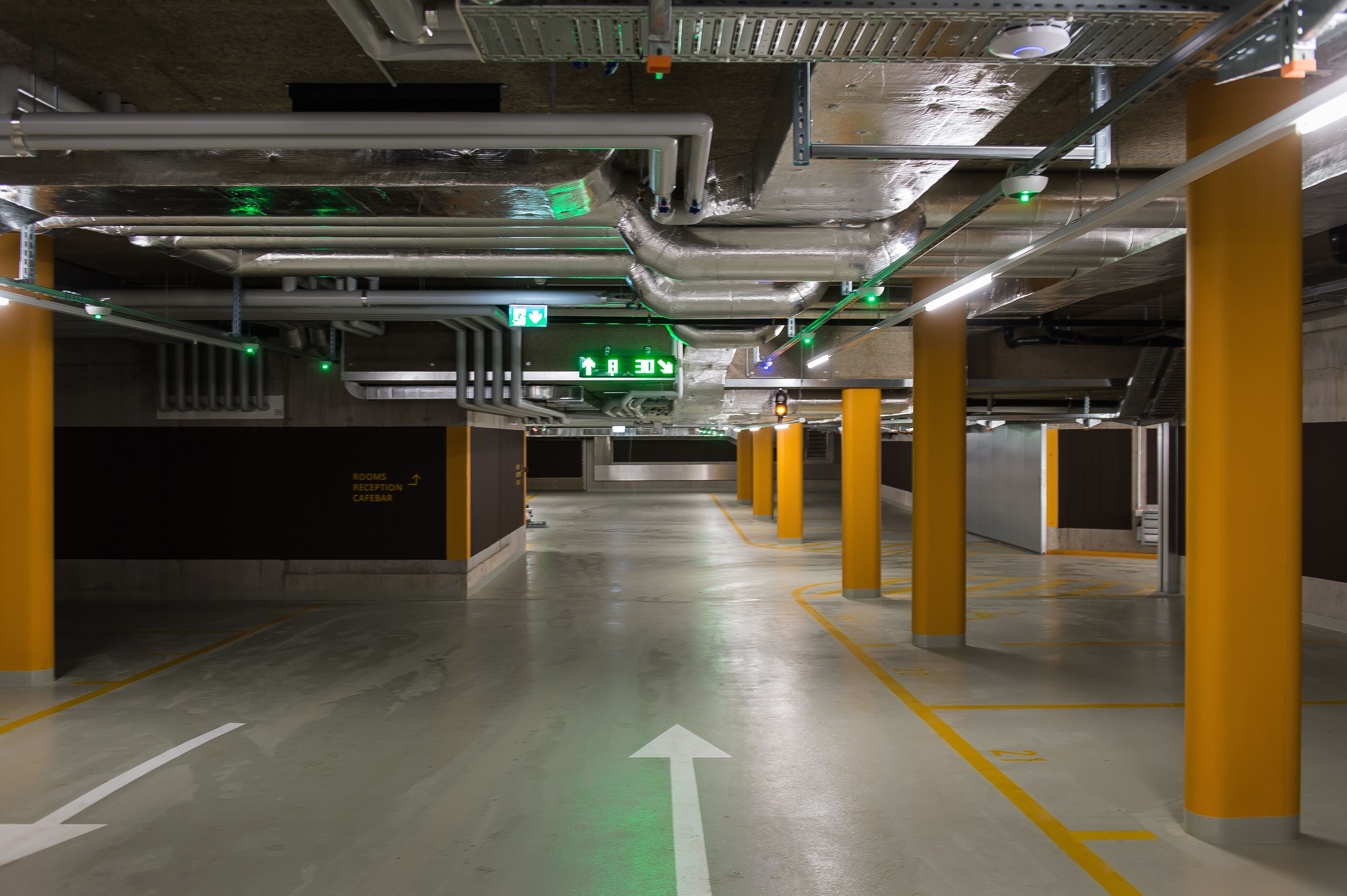 Un estacionamiento con flechas direccionales bien visibles en el suelo, pilares amarillos y señalización de salida verde.