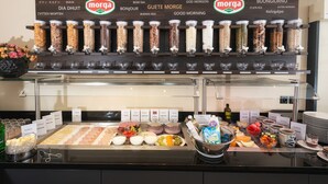 A breakfast buffet with labeled containers of granola and other cereals, a variety of fruits, and a selection of yogurts and cheeses.
