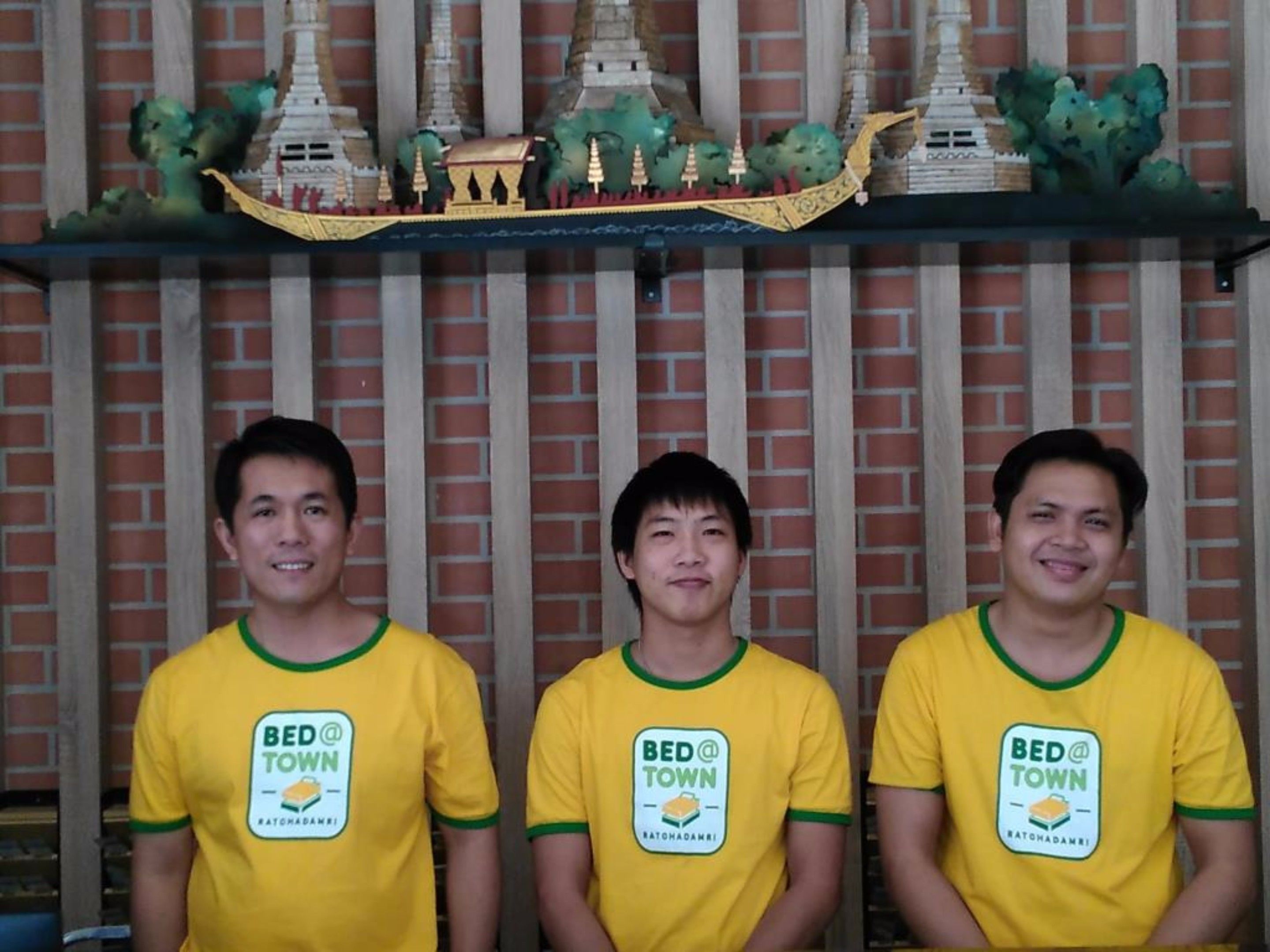 Three individuals wearing yellow t-shirts with 'BED @ TOWN' logos, standing in front of a brick wall with a decorative shelf above.