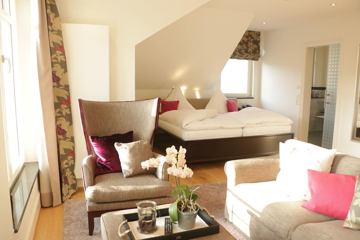 A bedroom with a bed, a chair, a sofa, a tray with a remote control, and a potted plant.