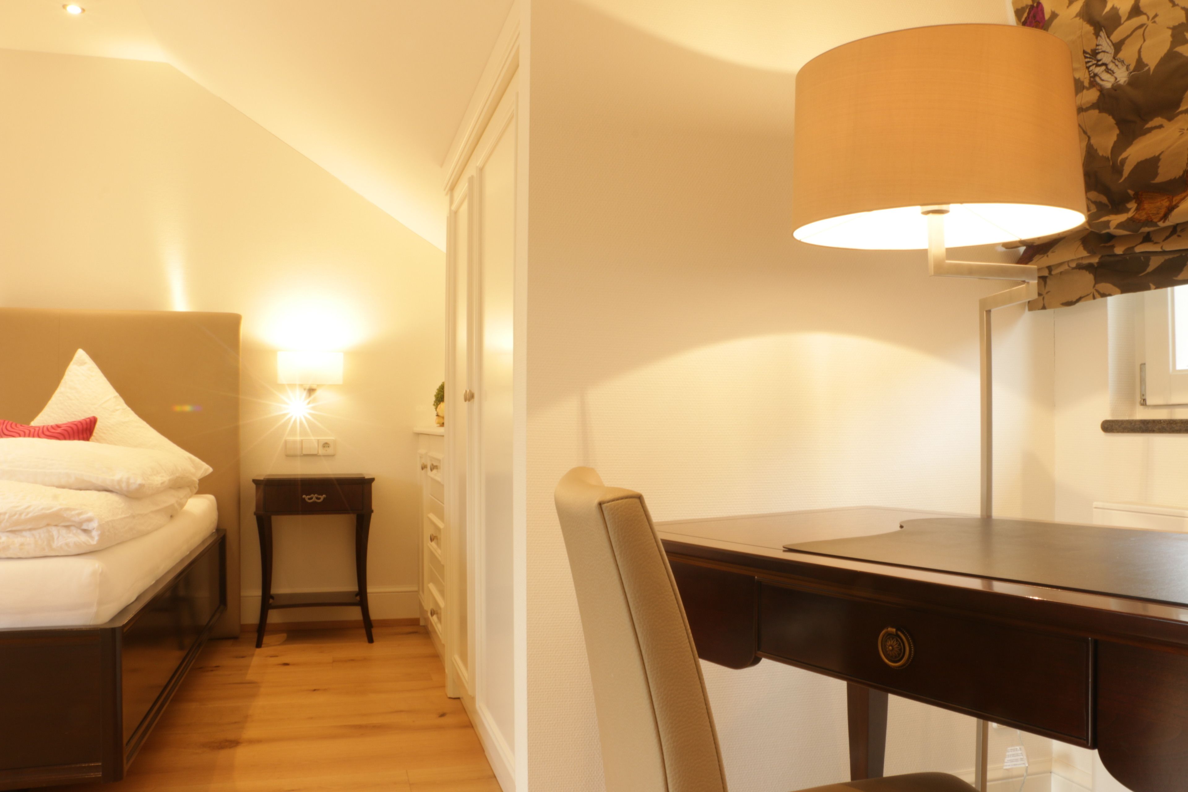 A hotel room with a bed, bedside table, lamp, desk, chair, and a window.