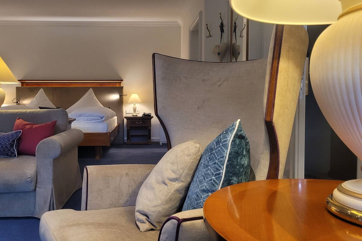 A hotel room with a sofa, armchair, bedside table with lamp, and a bed with pillows.