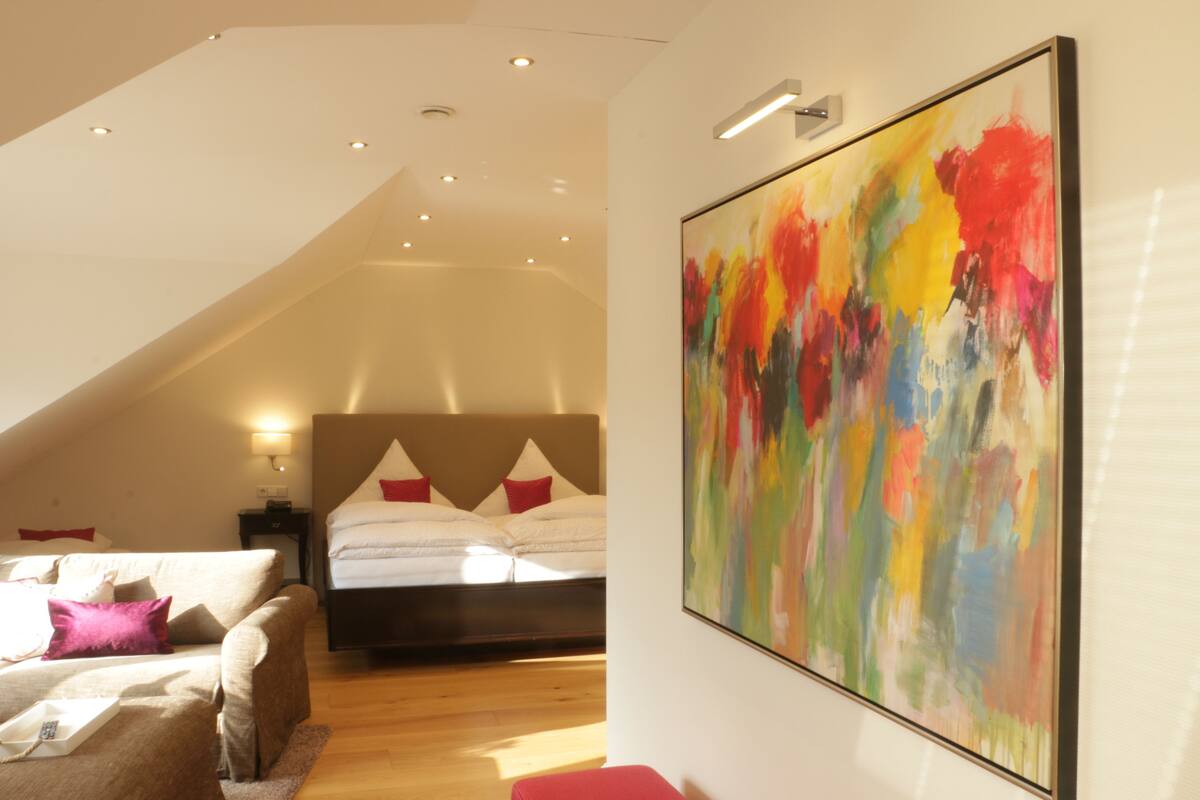 A modern bedroom with a sloped ceiling, a large bed with white linens, a colorful abstract painting, and a small bedside table with a lamp.