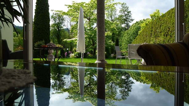 A view from a room with a glass table reflecting a garden with a white umbrella, chairs, and a hedge. - Baumanns Landhaus Durbach (Durbach)