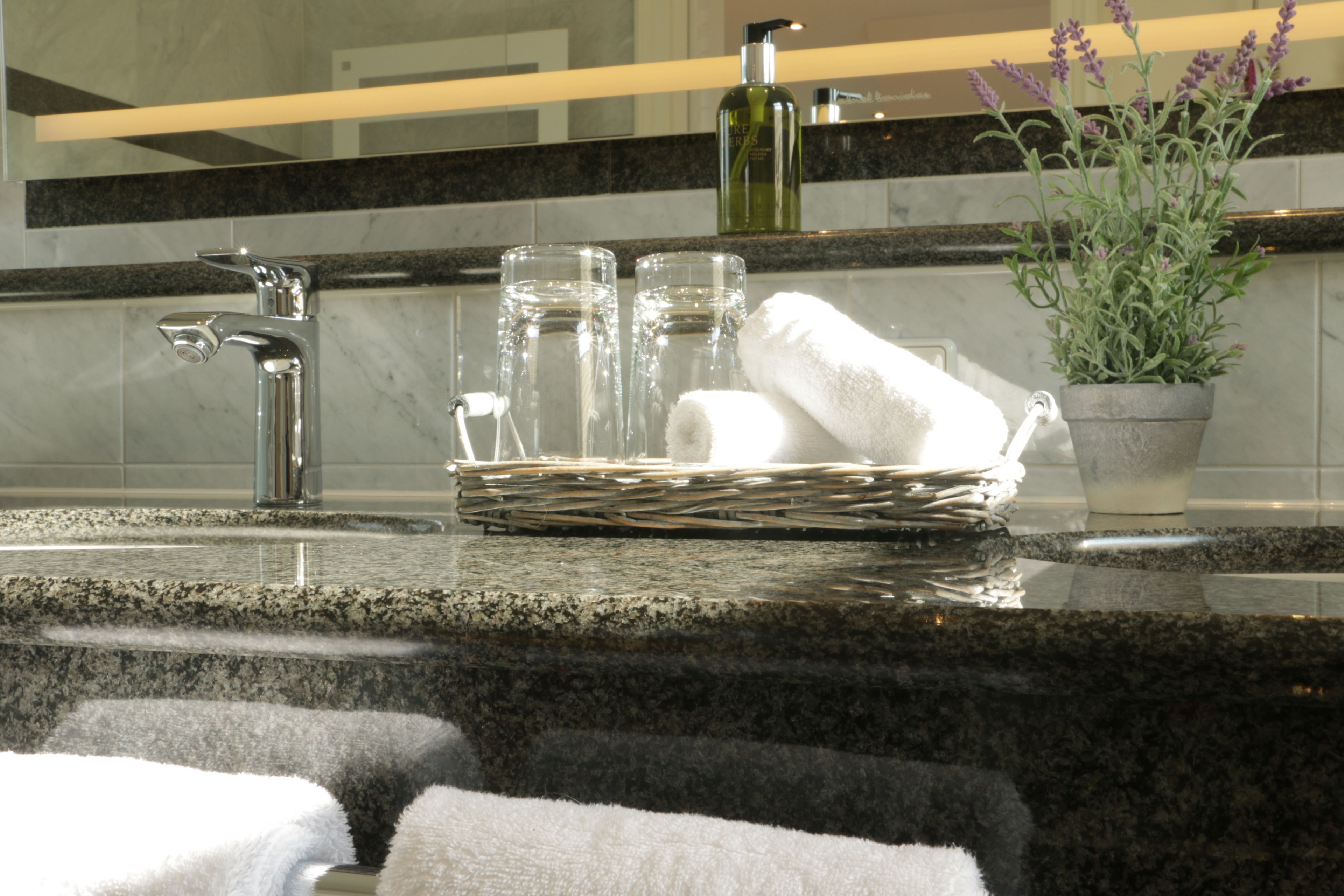 A bathroom with a granite countertop, a silver faucet, a glass of water, a bottle of lotion, a potted lavender plant, and two white towels.