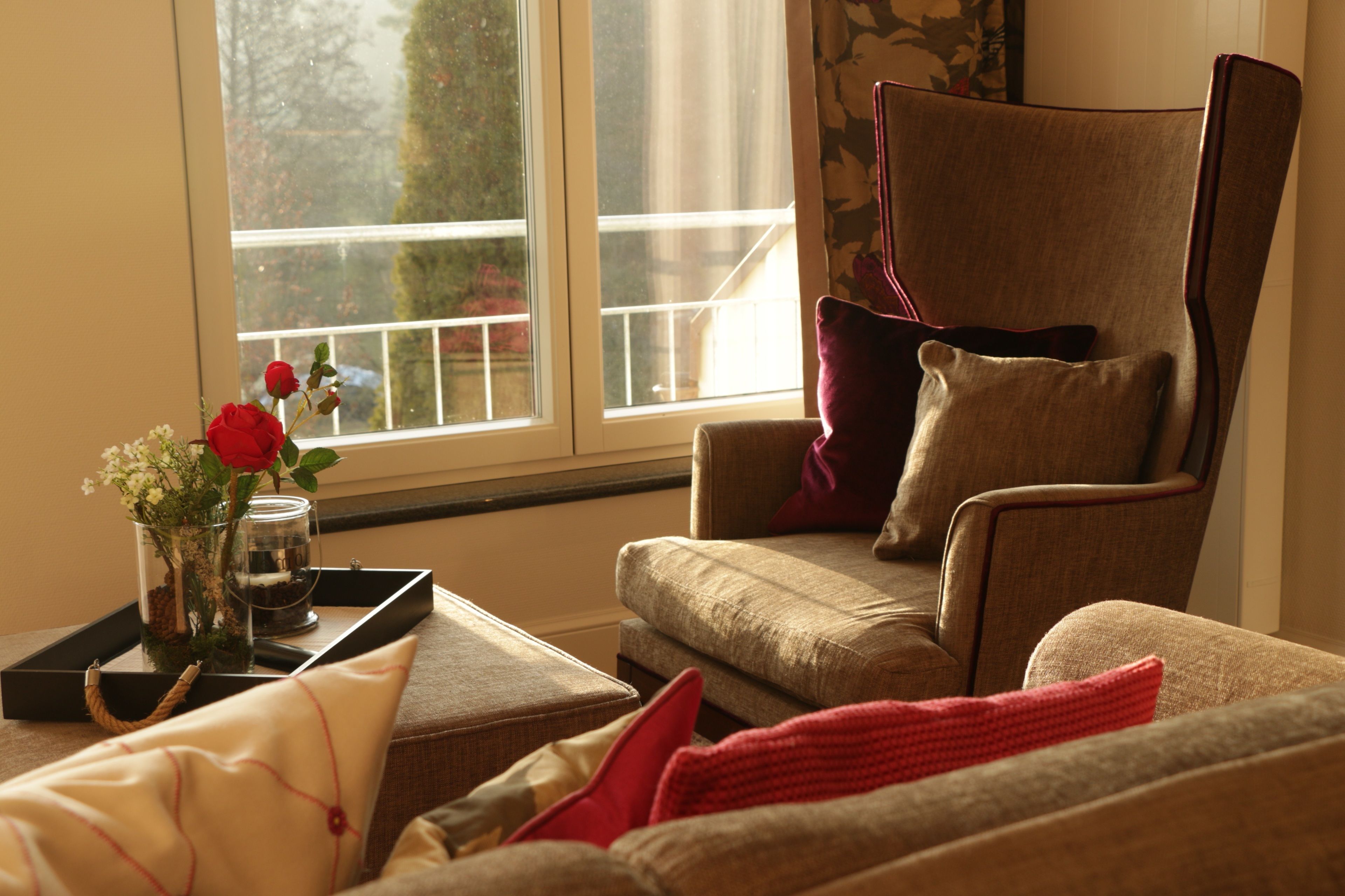 Een woonkamer met een grote fauteuil, een bijzettafeltje met een bloemetjesvaas en een raam met uitzicht op bomen.