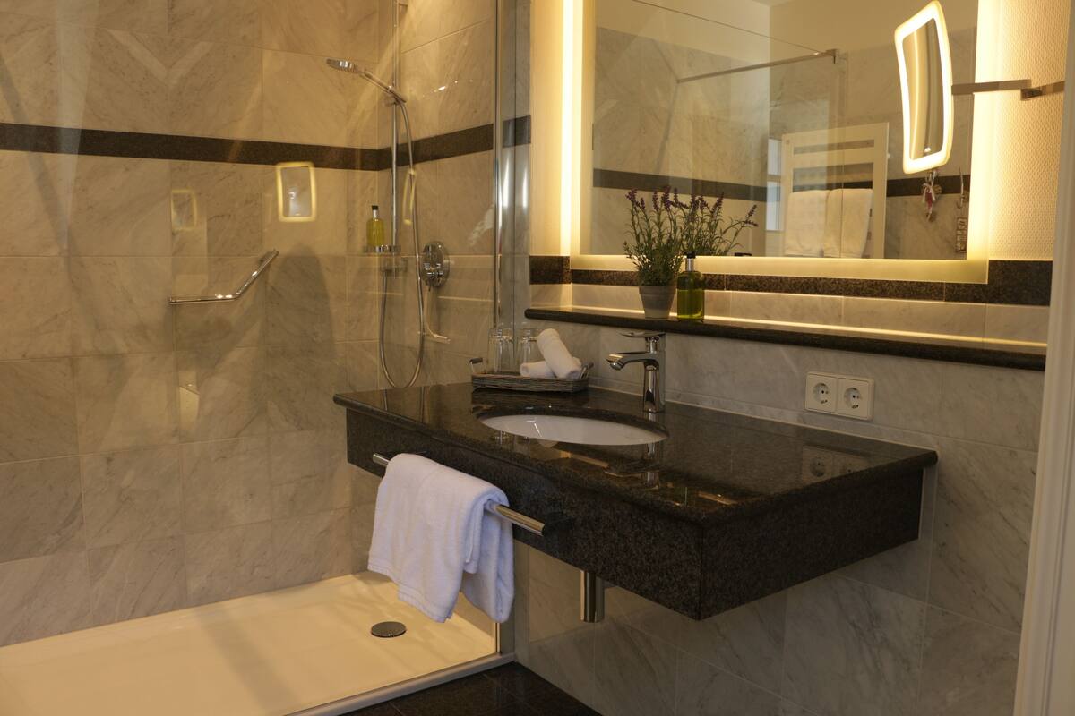 A modern bathroom with a glass-enclosed shower, a marble-tiled floor, a black granite countertop, a white sink, and a large mirror with integrated lighting.