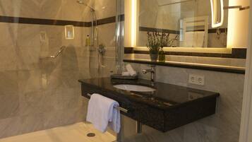 Un baño moderno con cabina de ducha de vidrio, suelo de mármol, encimera de granito negro, lavabo blanco y un espejo grande con iluminación integrada.