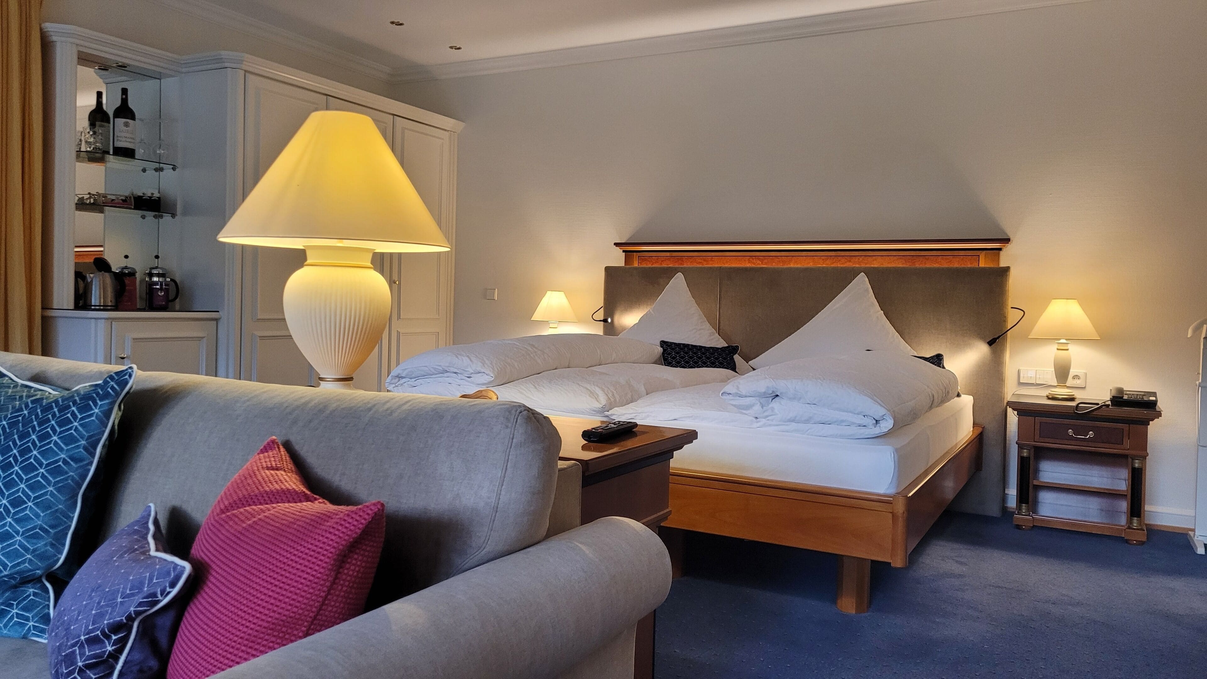 A hotel room with a bed, bedside tables, a sofa, a lamp, and a cabinet with bottles.