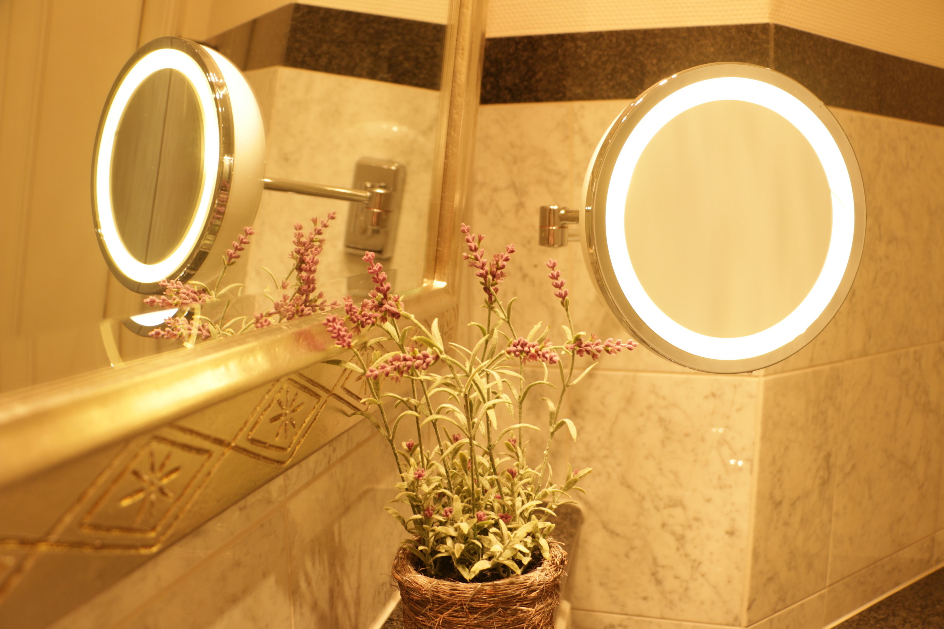 Un baño con dos espejos iluminados, una pared de mármol y una planta en maceta con flores moradas.