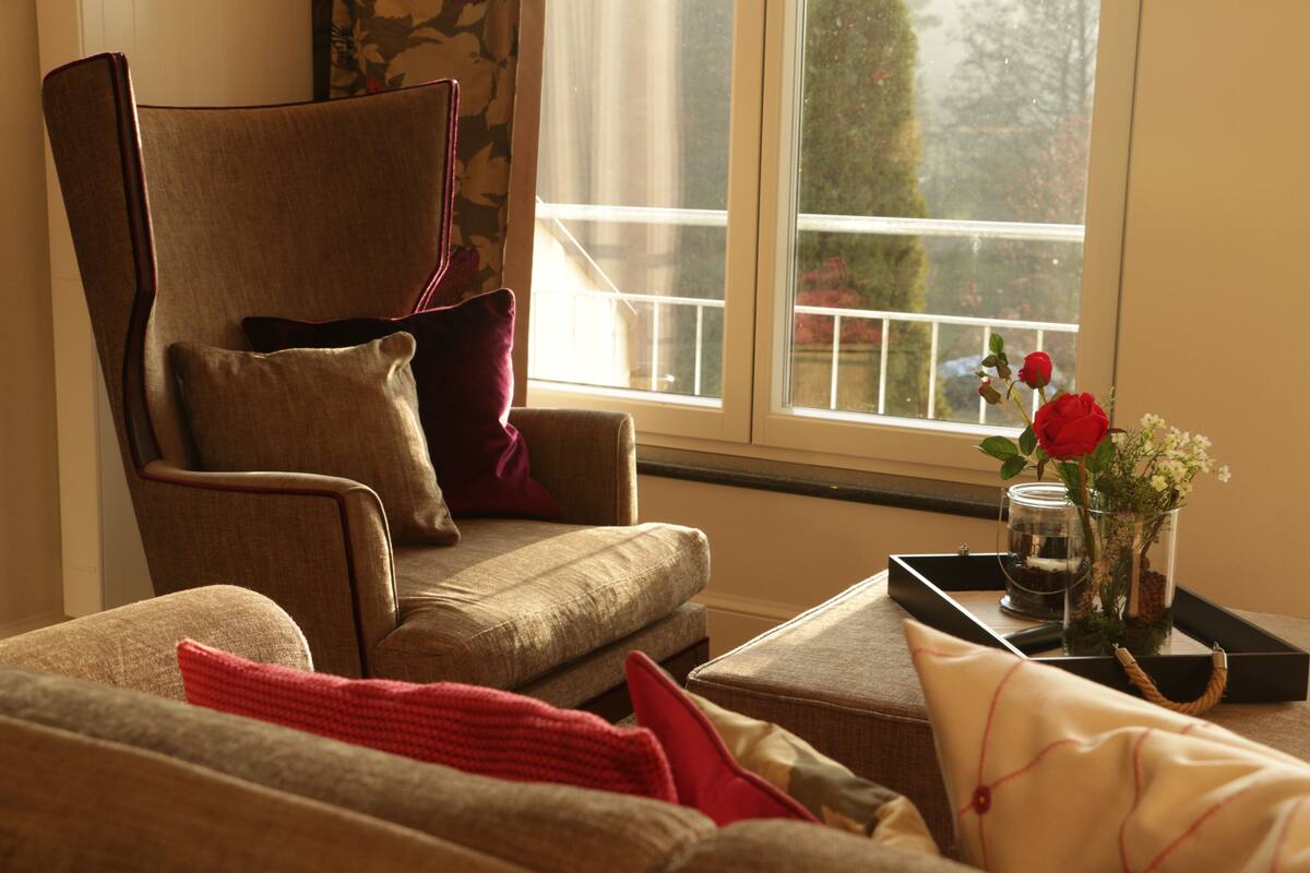 A cozy living room with a brown armchair, a small sofa, and a window with a view of a balcony.