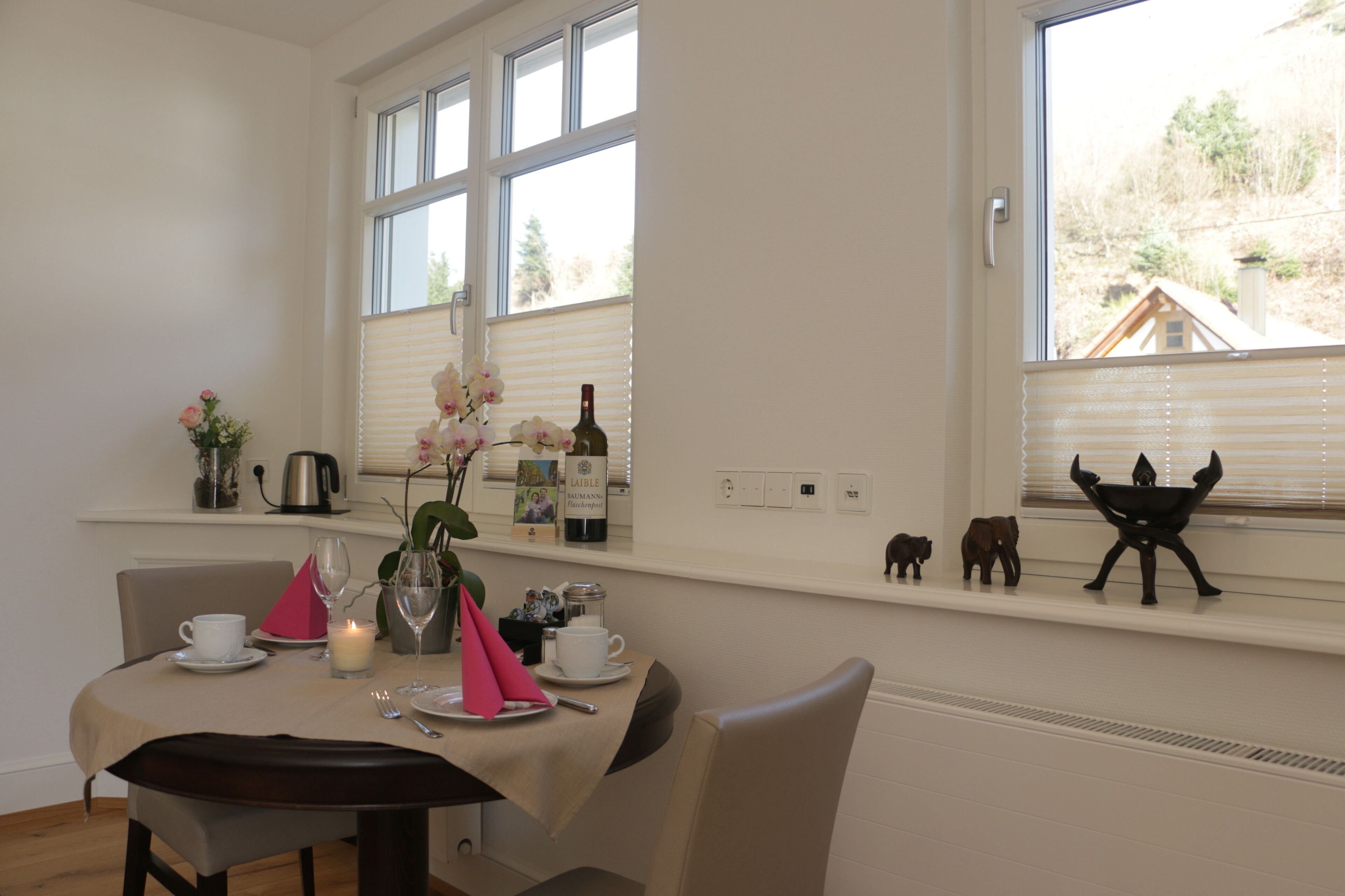 A dining area with a table set for two, a vase with flowers, a bottle of wine, and decorative items on the windowsill.