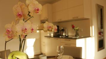 A dining area with a table set for two, a vase of orchids, and a kitchen in the background.