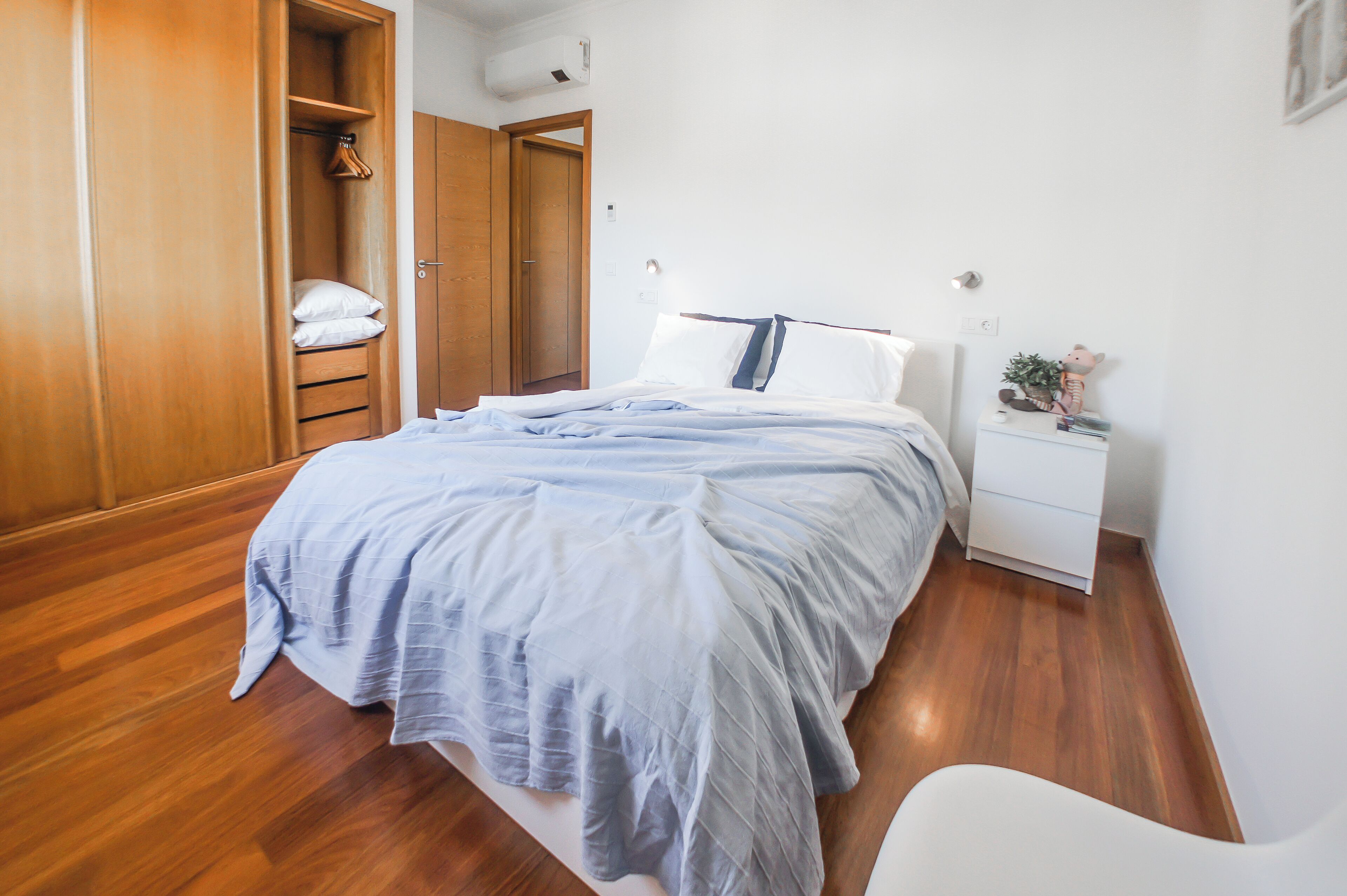 Ein Schlafzimmer mit einem Holzschrank, einem Bett mit blauer Bettdecke, einem weißen Nachttisch mit Pflanze und einem weißen Stuhl.