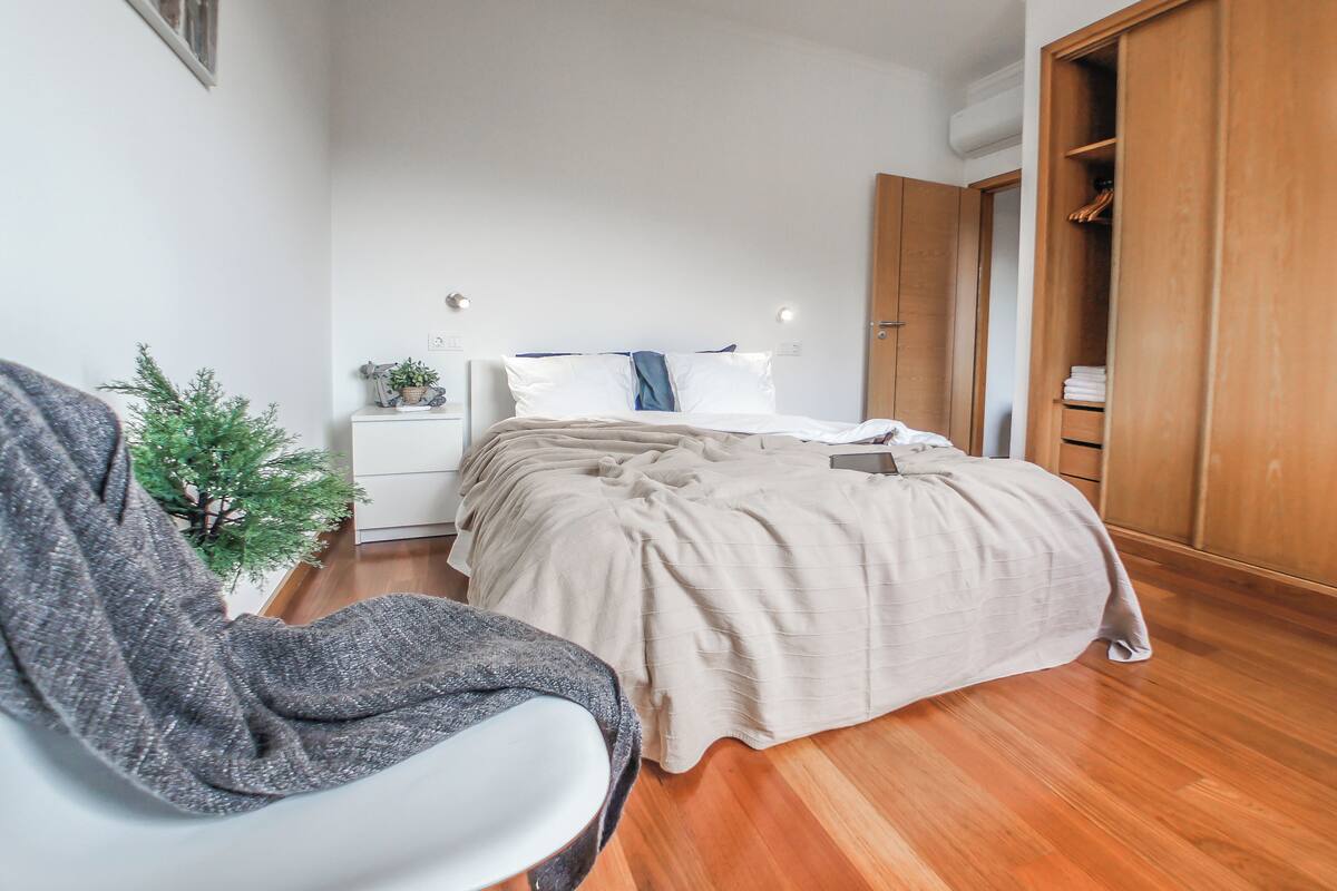 A bedroom with a bed, a chair, a small plant, and a wooden wardrobe.