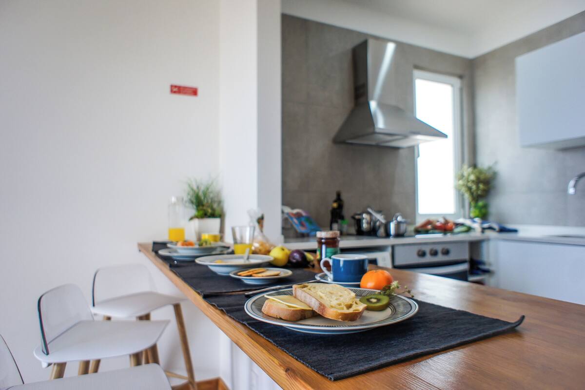 A modern kitchen with a dining table set for breakfast, including plates of toast, fruit, and a cup of coffee.