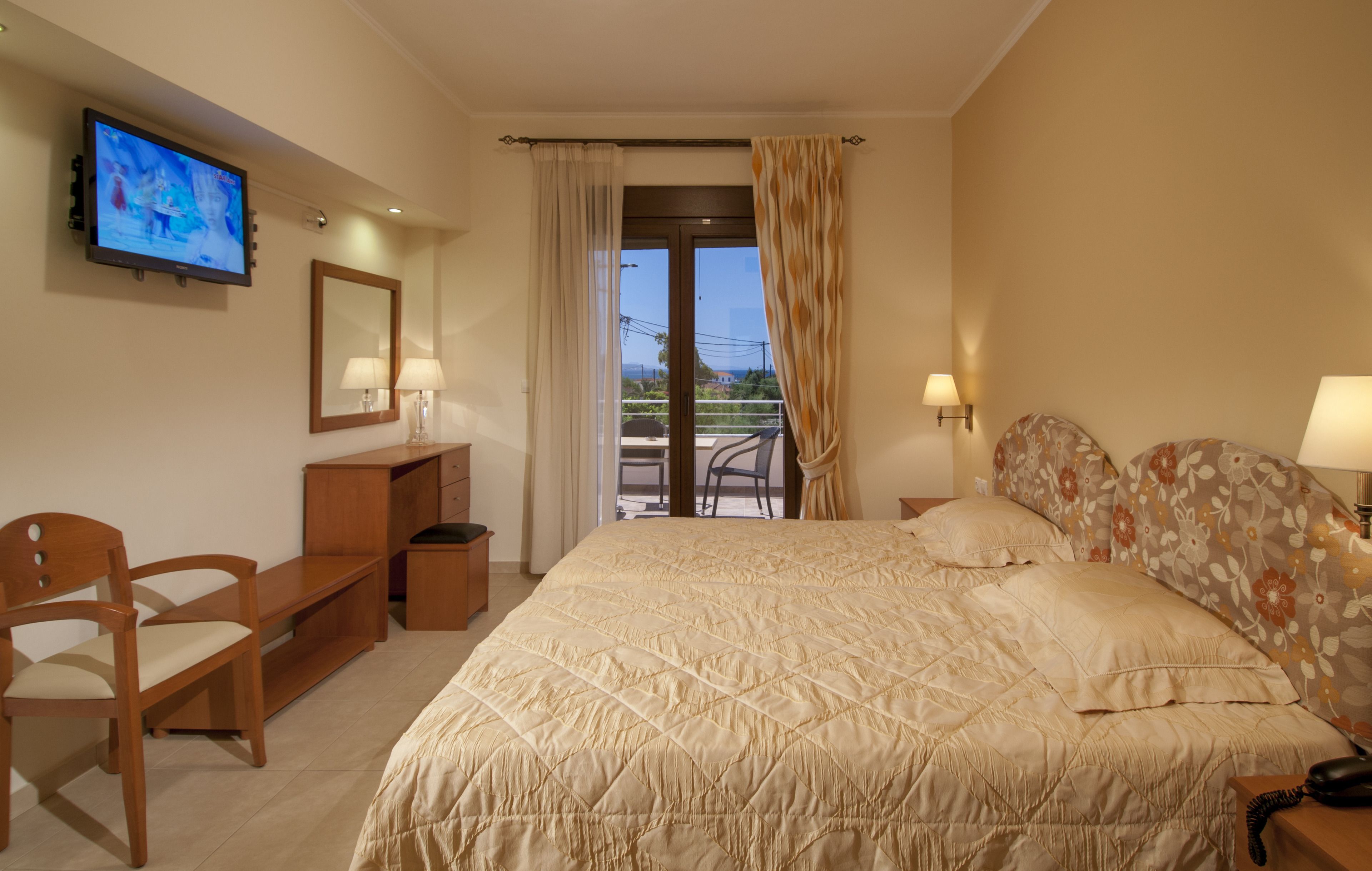 A hotel room with a bed, a TV, a desk, a chair, and a balcony with curtains.