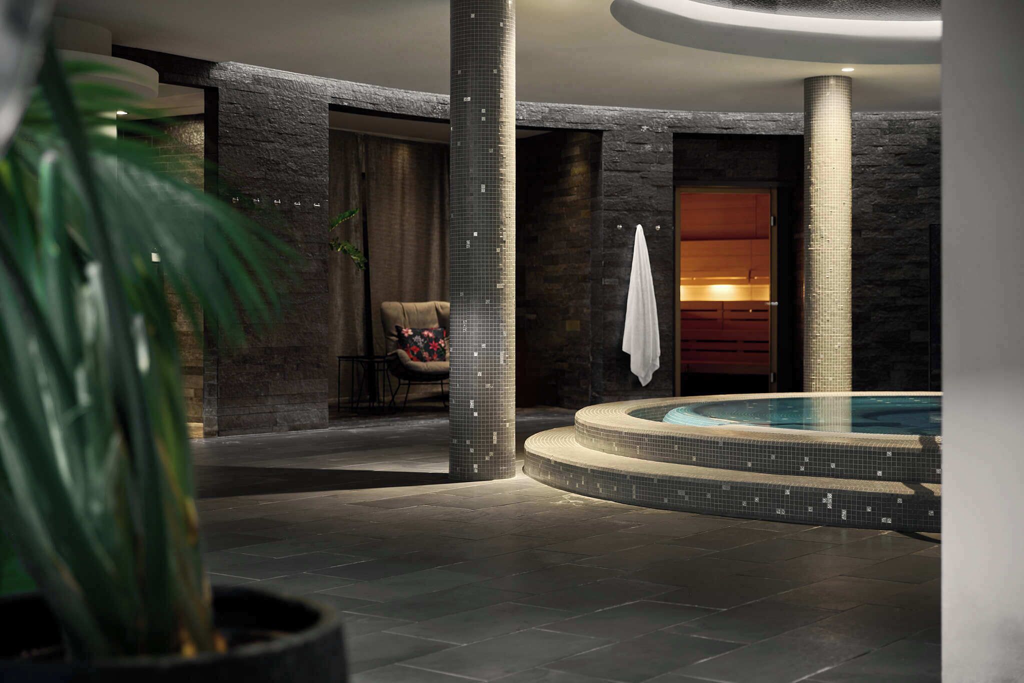 Ein moderner Wellnessbereich mit einer runden Whirlwanne, Steinwänden und einer Sitzgelegenheit mit Stuhl und Handtuch.