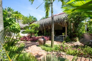 Un jardín tropical con una estructura de techo de paja, abundante vegetación y un sendero de piedra.