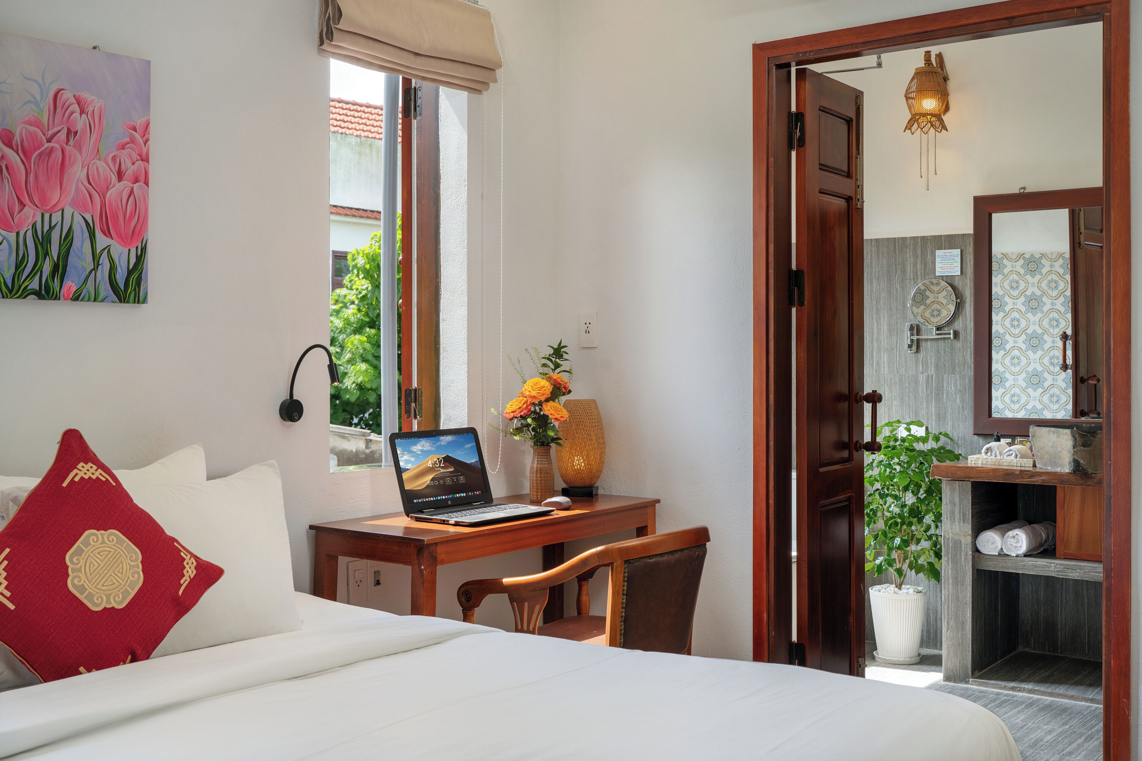 Un dormitorio con cama, escritorio con computadora portátil, silla, almohada roja, florero con flores y puerta que da a un baño.