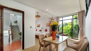 A modern living room with a glass dining table, beige chairs, a large window with a view of greenery, a staircase, and a bed with a white headboard. - Red Flower Cottages Homestay (Da Nang)