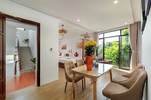 Una sala moderna con una mesa de comedor de vidrio, sillas color beige, un ventanal con vistas a la vegetación, una escalera y una cama con cabecera blanca.