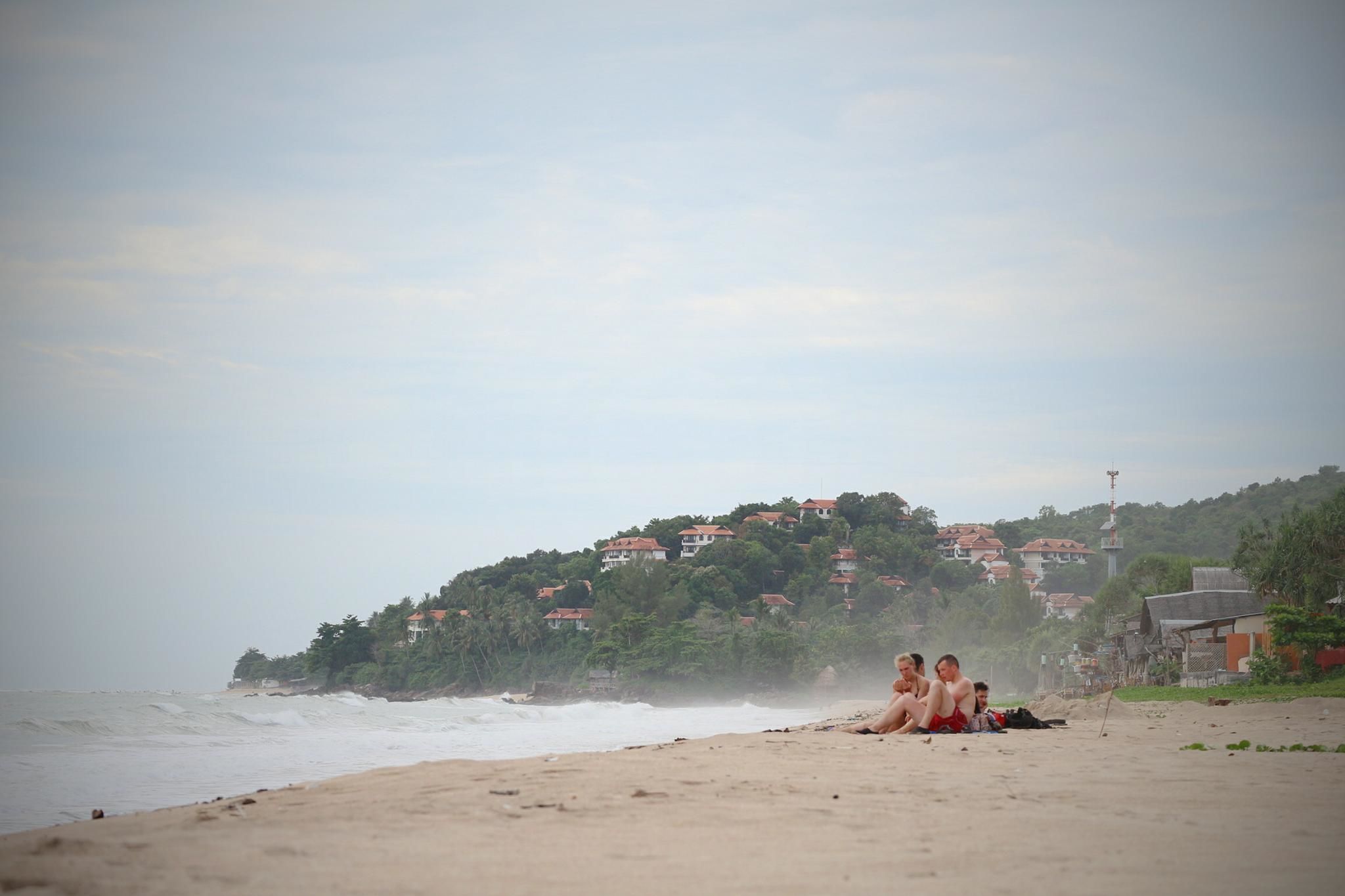 A beach with a sandy shore, a few people sitting on the sand, and houses on a hill in the background.