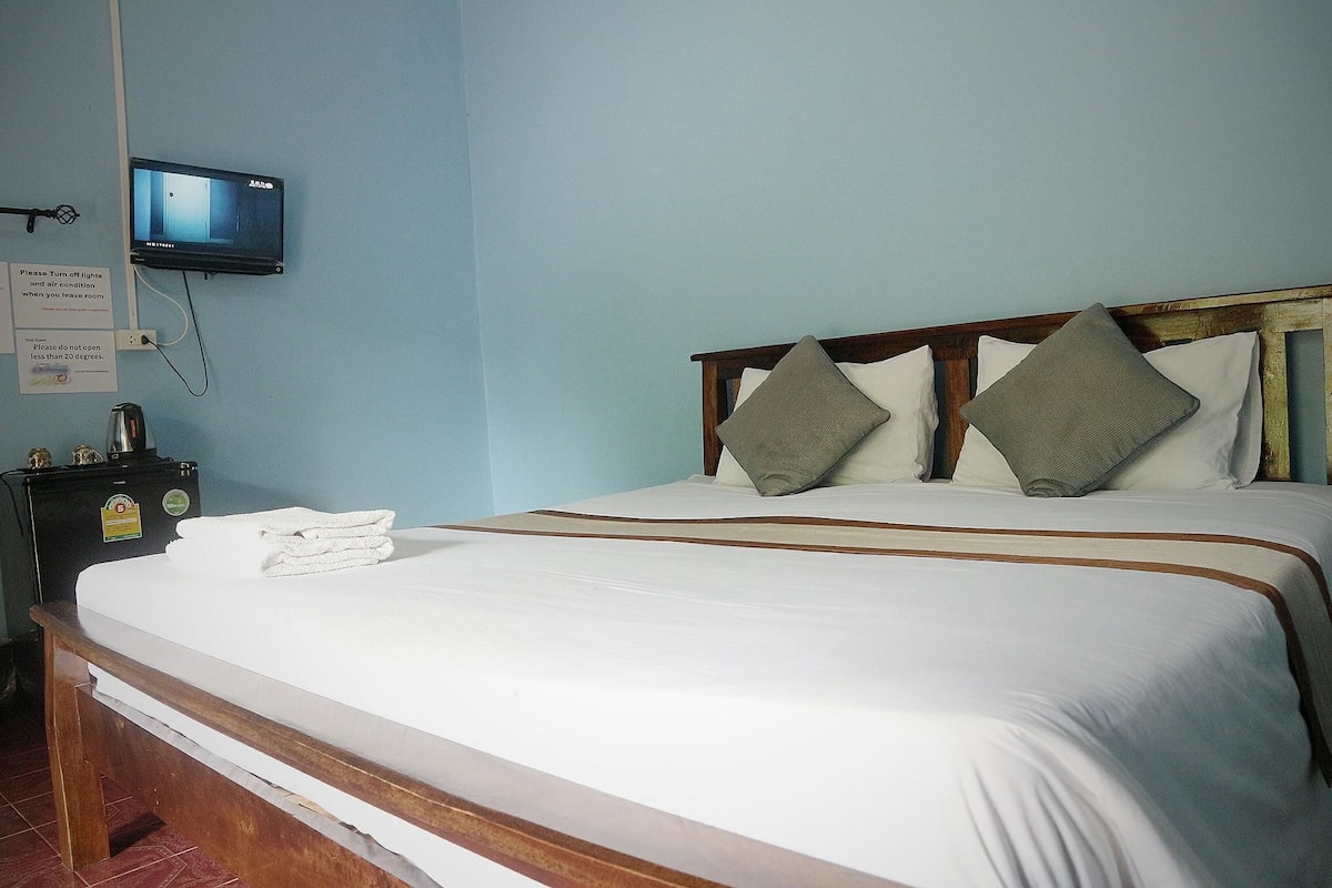 Uma cama bem arrumada com cabeceira de madeira, televisão fixada na parede e uma pequena mesa com abajur e um aviso.