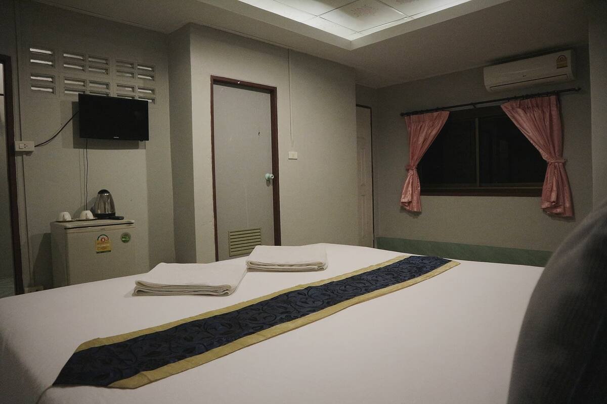 Quarto de hotel com cama, secretária com chaleira, televisão fixada na parede e janela com cortinas.