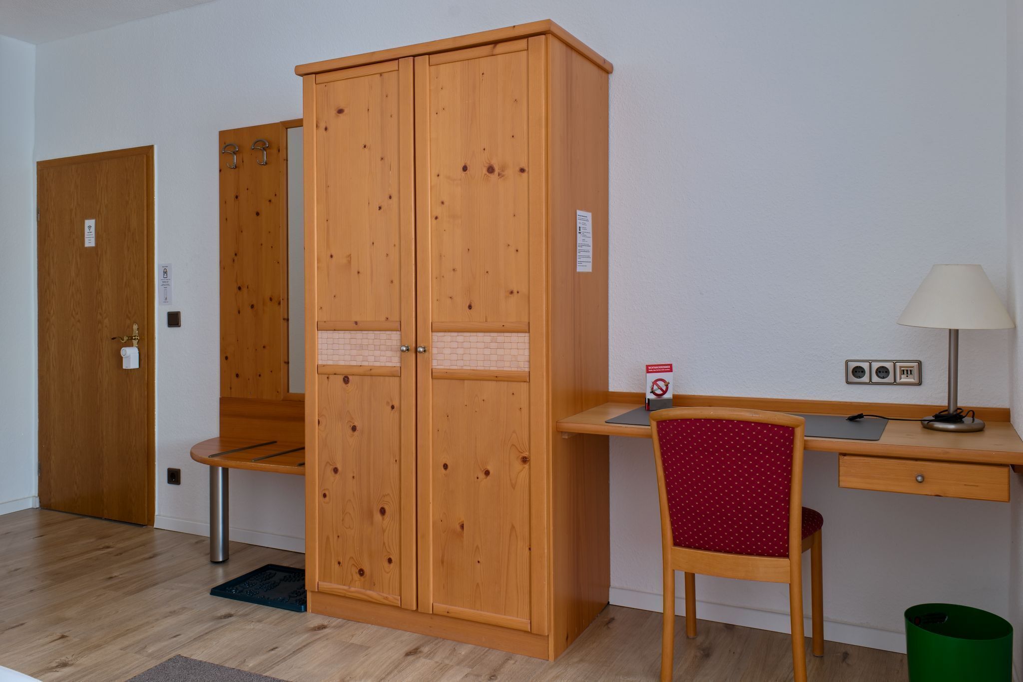 A room with a wooden wardrobe, a desk with a chair, a lamp, and a trash bin.