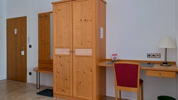 A room with a wooden wardrobe, a desk with a chair, a lamp, and a trash bin.