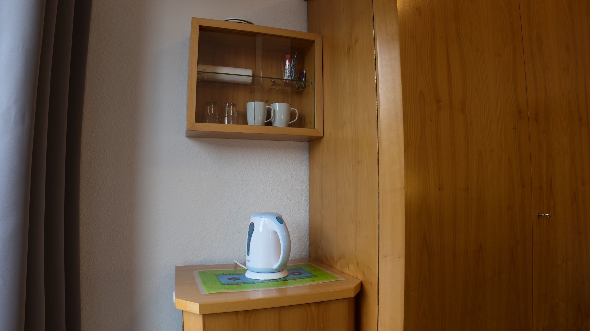 A wooden cabinet with cups and a kettle on a small table.