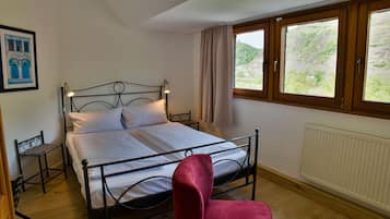 A single bed with a headboard, a bedside table, a chair, a window with curtains, and a radiator.