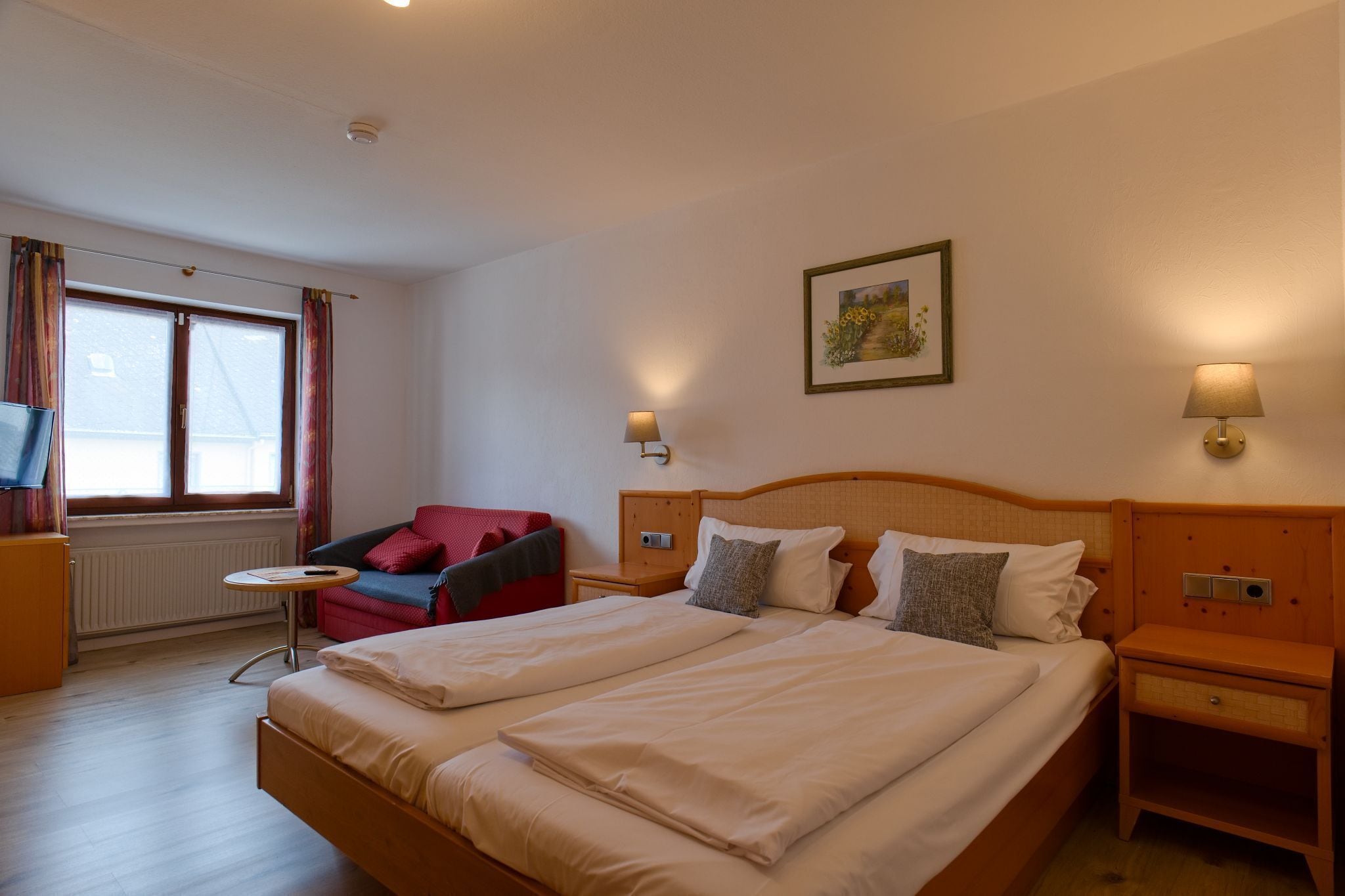 Ein Doppelbett mit zwei Nachttischen, ein kleiner runder Tisch, ein roter Sessel und ein Fenster mit Vorhängen.