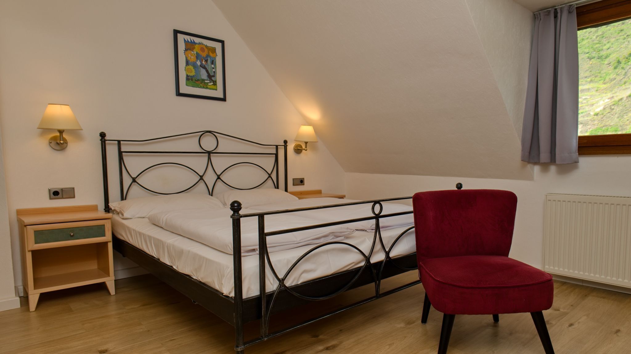 A single bed with a metal frame, two bedside tables, a red chair, a framed picture on the wall, and a window with curtains.