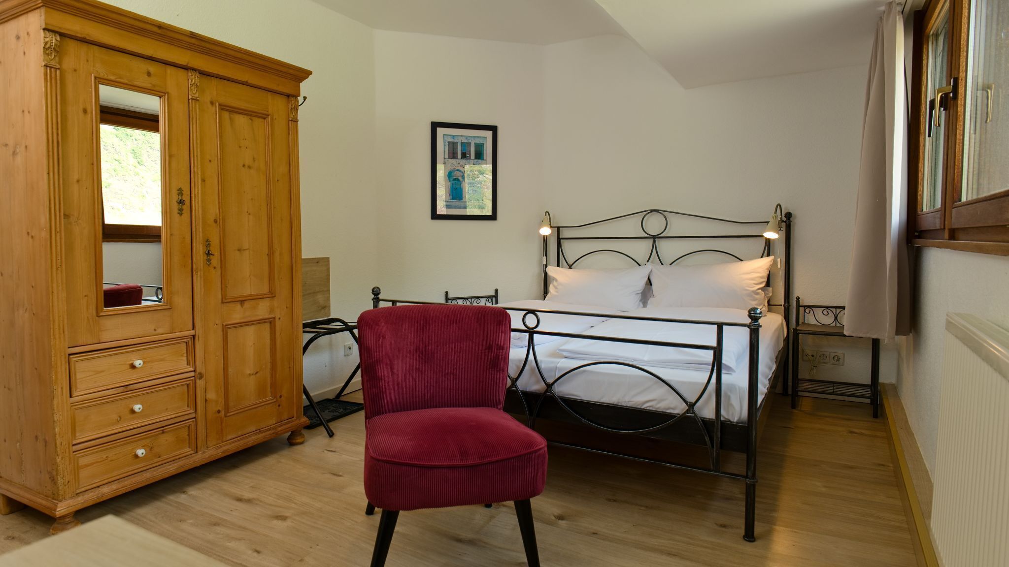 A bedroom with a wooden wardrobe, a red chair, a metal bed frame, and a framed picture on the wall.