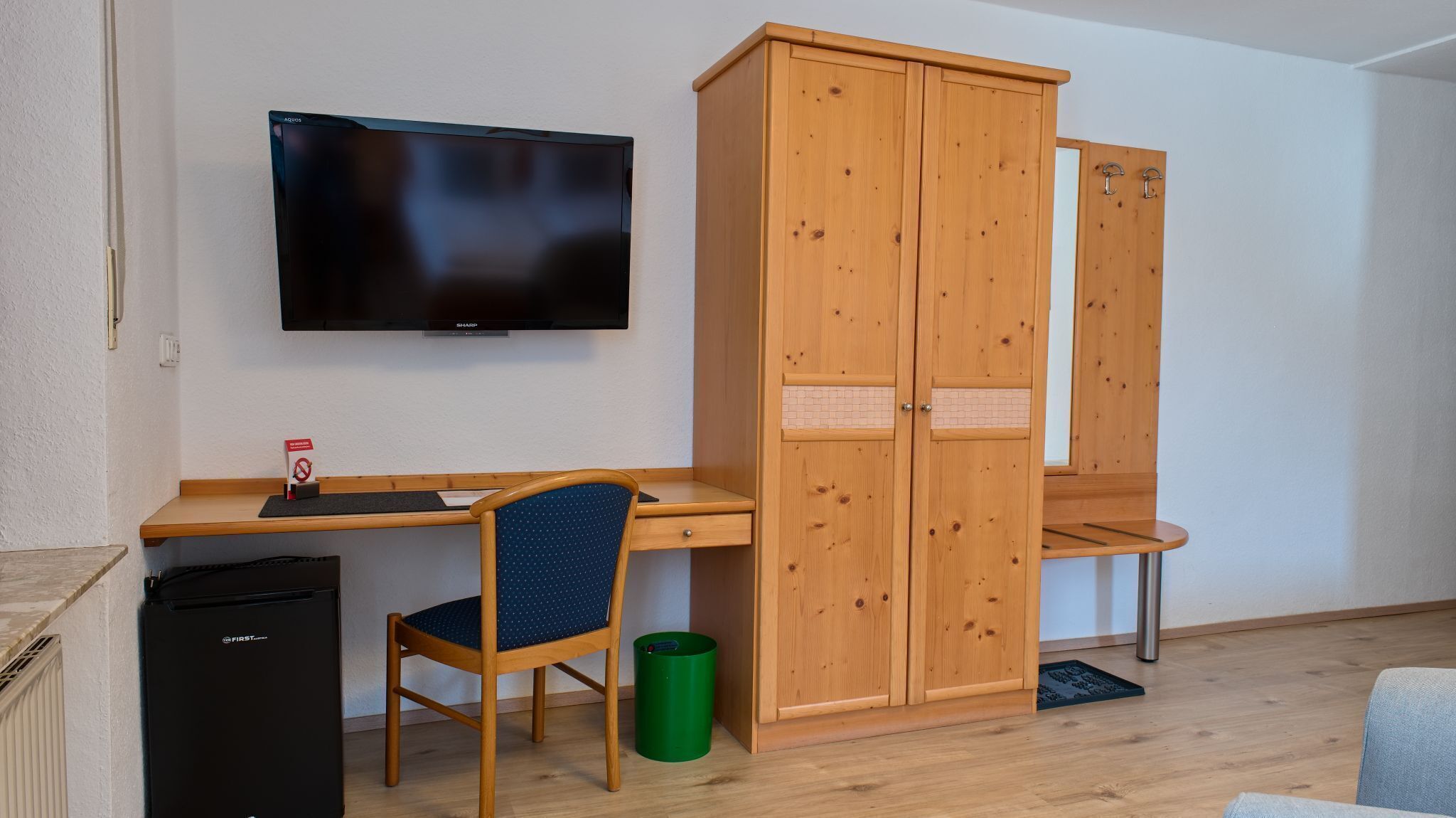 Una habitación con escritorio de madera, una silla, televisor, armario y cubo de basura.