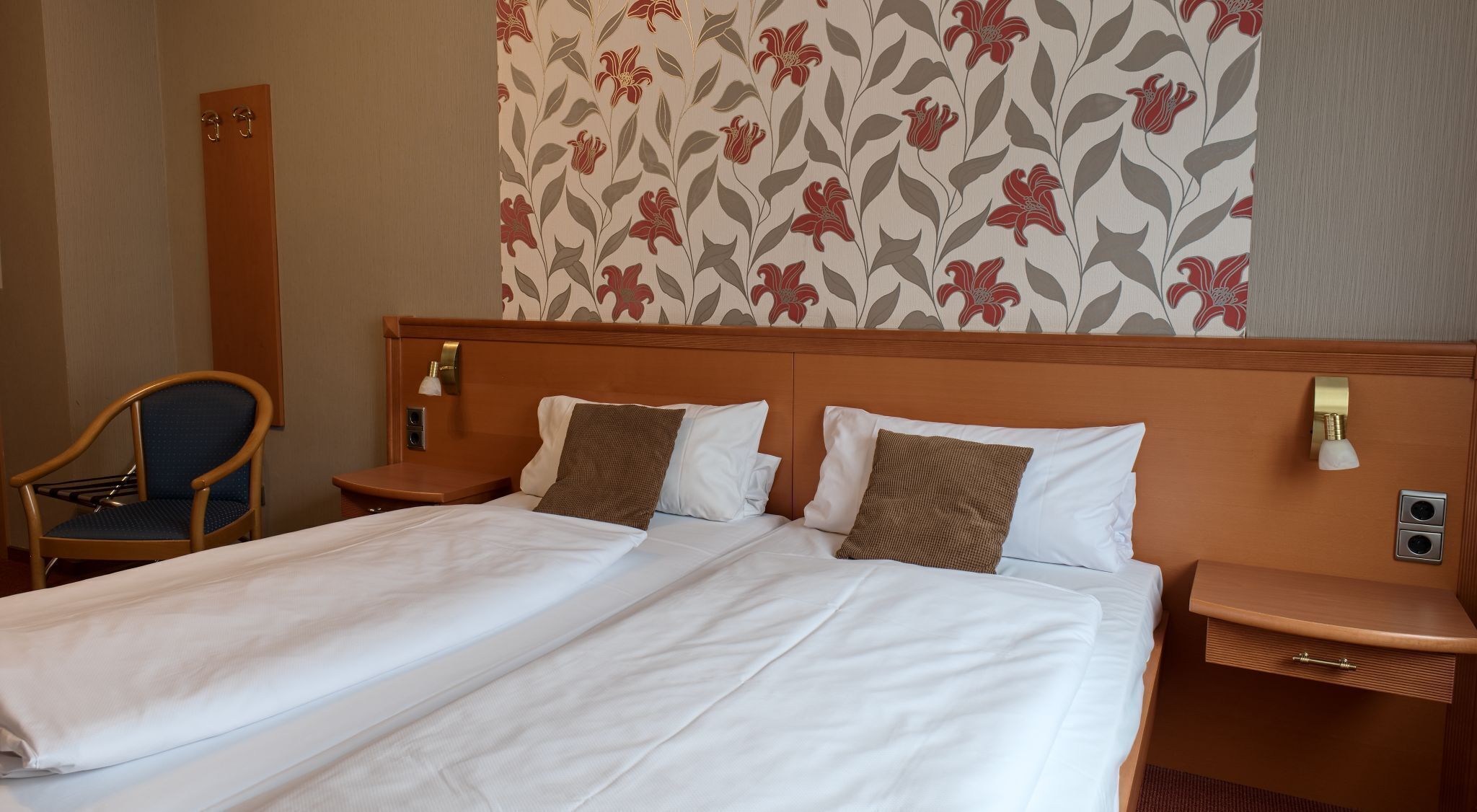 Une chambre d’hôtel avec un lit double, deux tables de chevet, une chaise et un papier peint à motifs floraux.