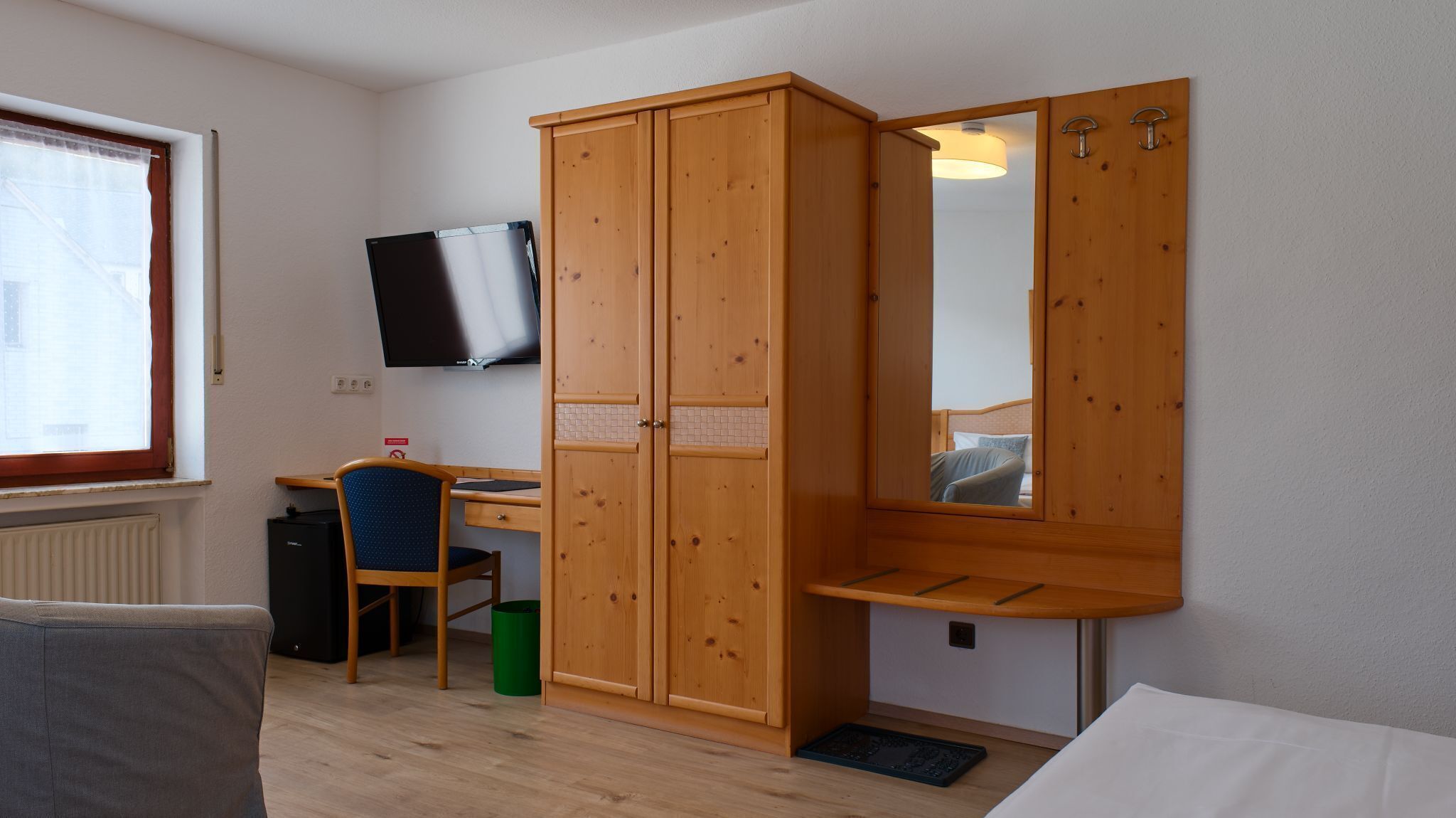 Una habitación con un armario de madera, un escritorio con una silla, un televisor fijado a la pared y una cama.