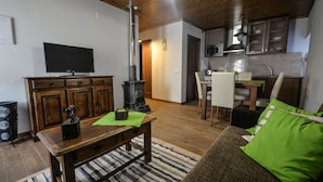 A living room with a television, wooden furniture, a green cushion, and a kitchen area with a microwave and cabinets.