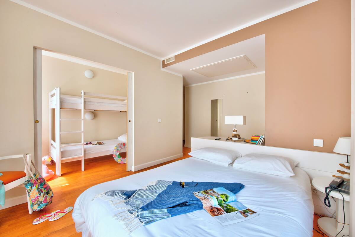 A hotel room with a bunk bed, a large bed with a blue blanket, a bedside table with a lamp, and a bathroom with a mirror and towel rack.