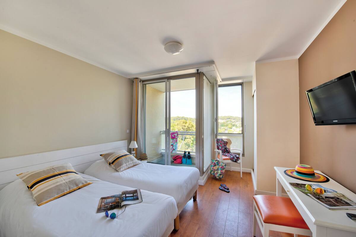 A hotel room with two beds, a balcony, a TV, and a desk with a hat and sunglasses.