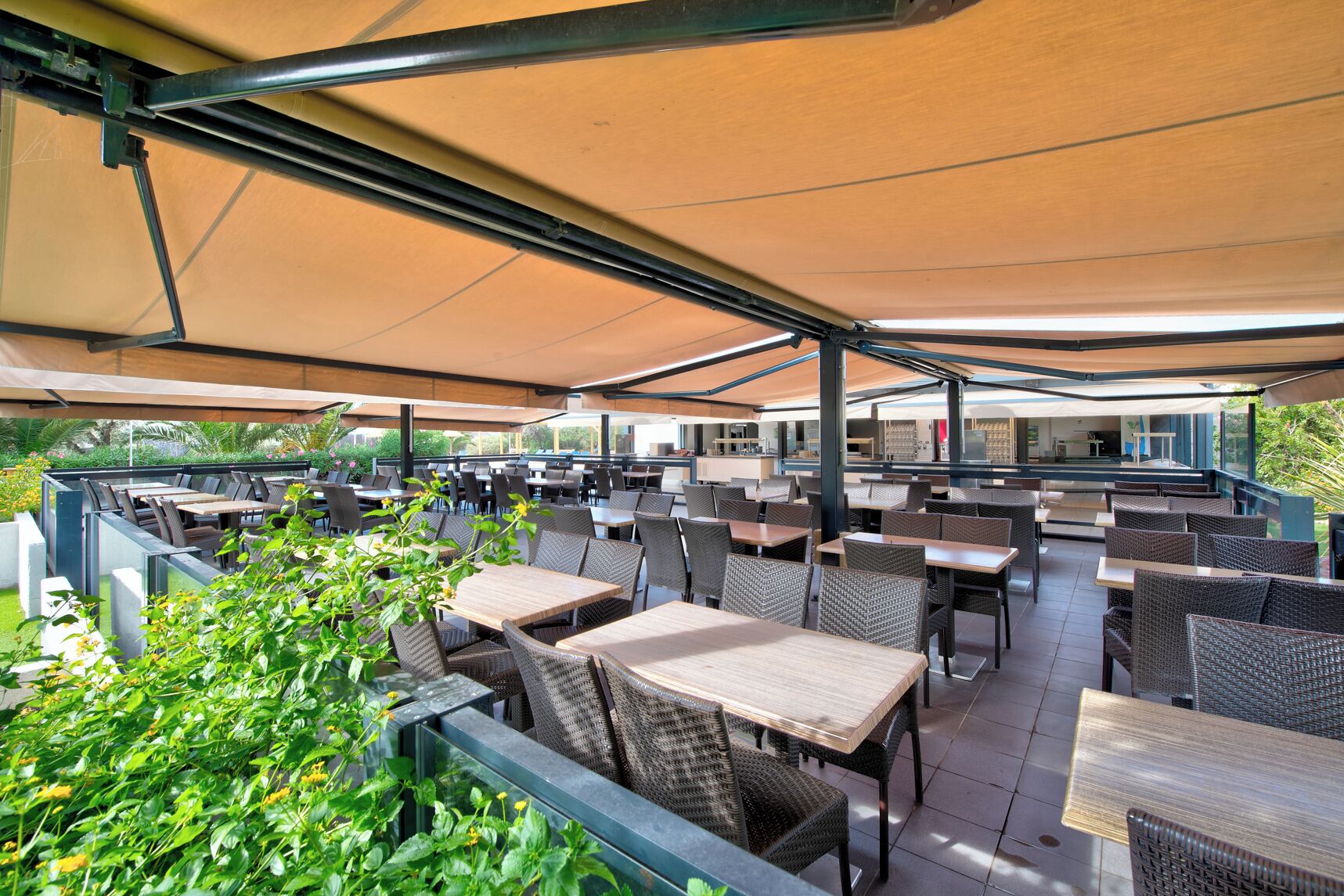 An outdoor patio with multiple tables and chairs, surrounded by greenery and covered by a large canopy.