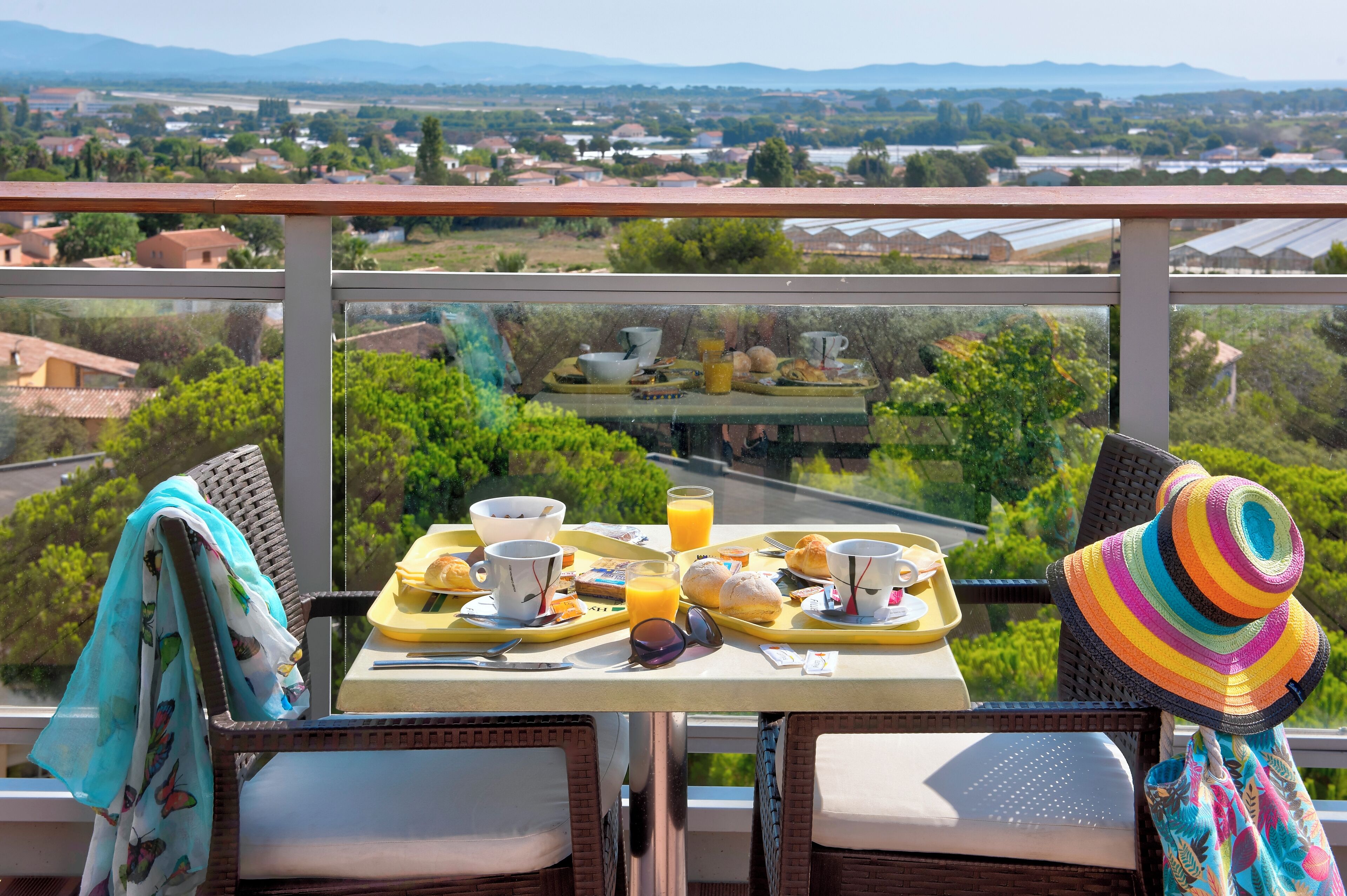 Un balcon avec une table dressée pour le petit-déjeuner, offrant une vue panoramique sur un paysage pittoresque, avec des maisons et de la verdure.