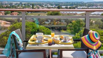 Un balcon avec une table dressée pour le petit-déjeuner, offrant une vue panoramique sur un paysage pittoresque, avec des maisons et de la verdure.