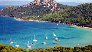 A coastal landscape with sailboats on clear blue waters, surrounded by dense green hills and a rocky mountain in the background.