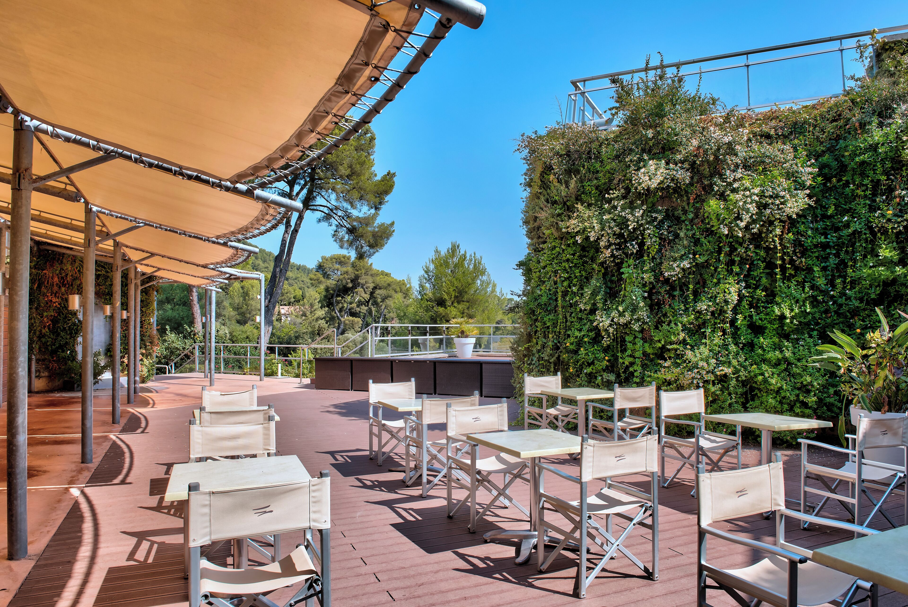 Een terras buiten met tafels, stoelen, een luifel en uitzicht op groen.