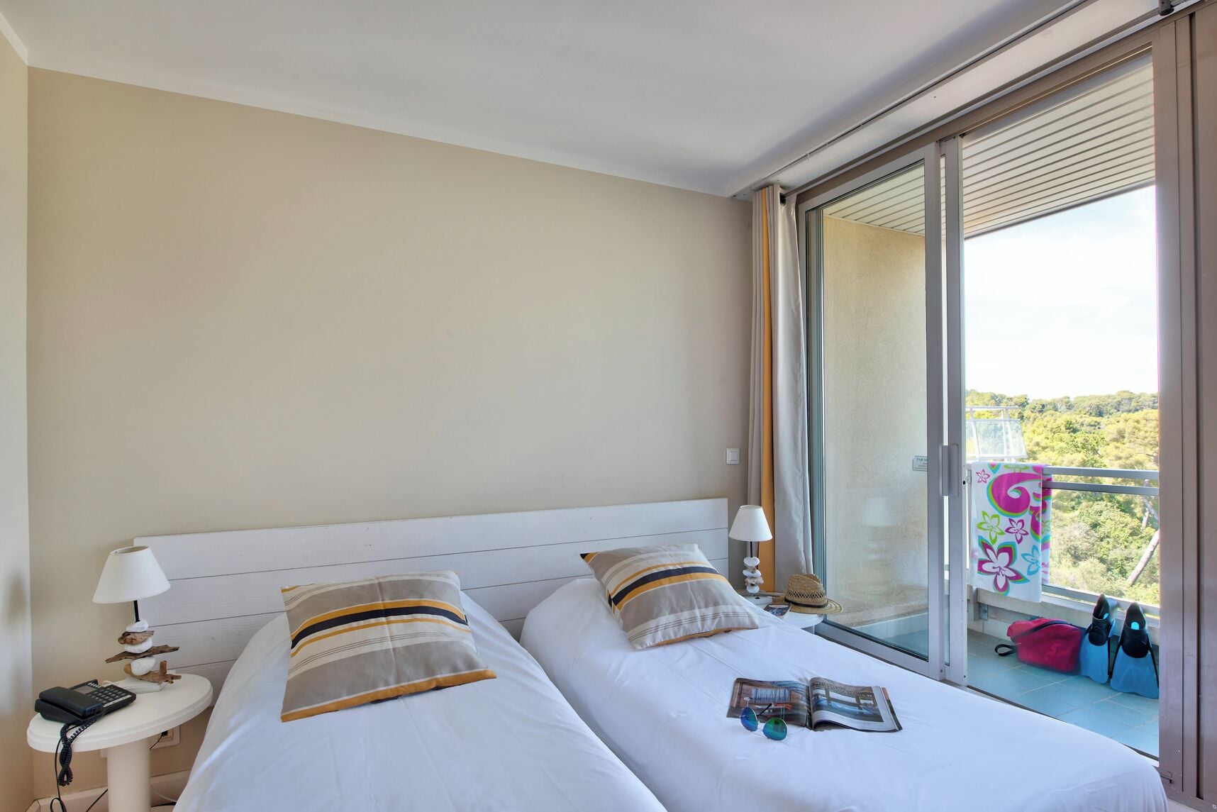 Une chambre d’hôtel avec deux lits, une petite table avec une lampe, une fenêtre donnant sur un espace verdoyant et un balcon avec une chaise et des sacs.